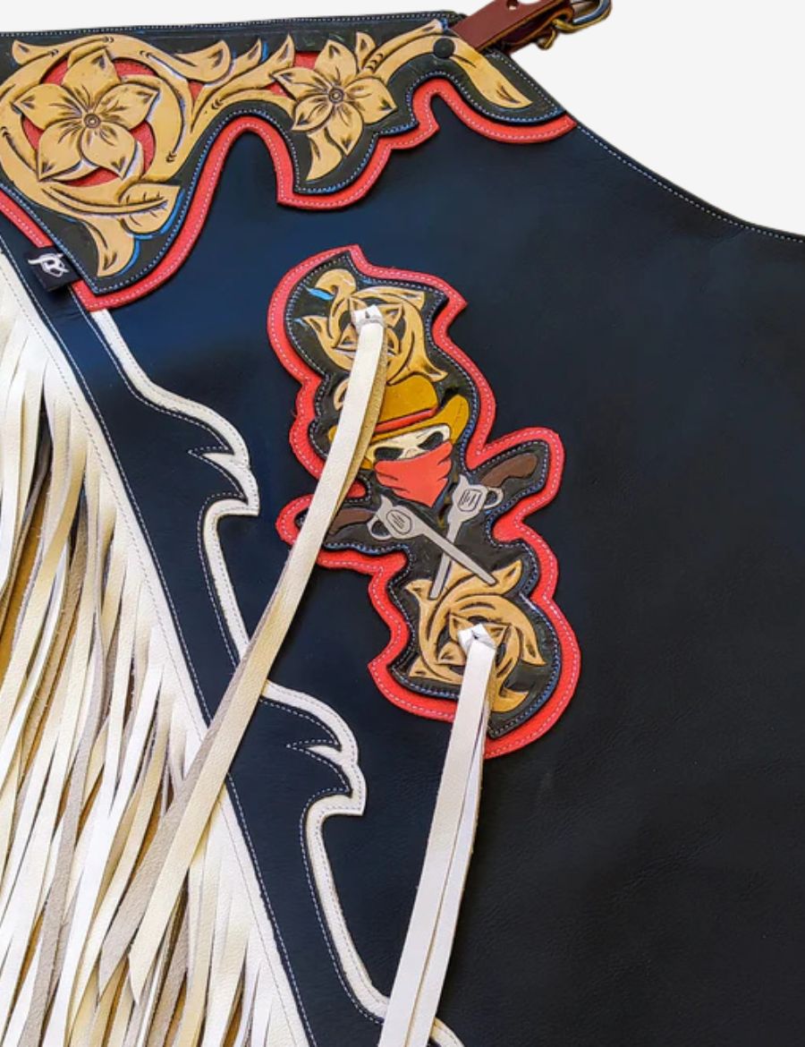 Detailed view of black rodeo chaps featuring cowboy graphic artwork, white lace straps, and western fringe accents.