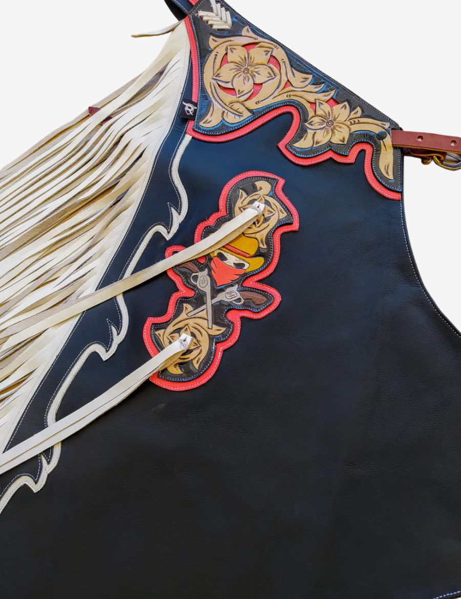 Close-up of black rodeo chaps showcasing floral tooled yoke, red piping, and handcrafted western rodeo detailing.