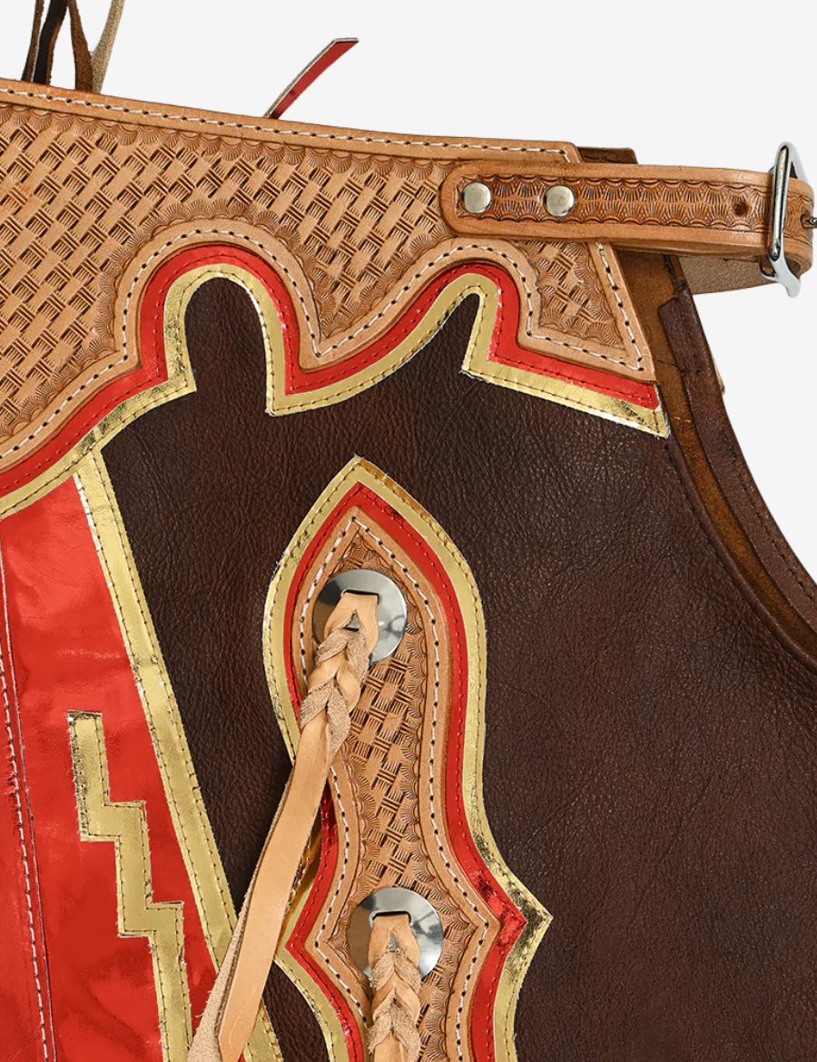 Tooled yoke detail of brown leather rodeo chaps featuring metallic orange fringe and western floral carving.