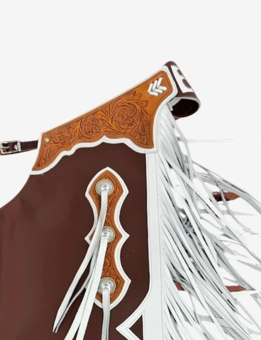 Close-up tooling detail on brown and white bull riding chaps featuring floral carved leather and western rodeo accents.