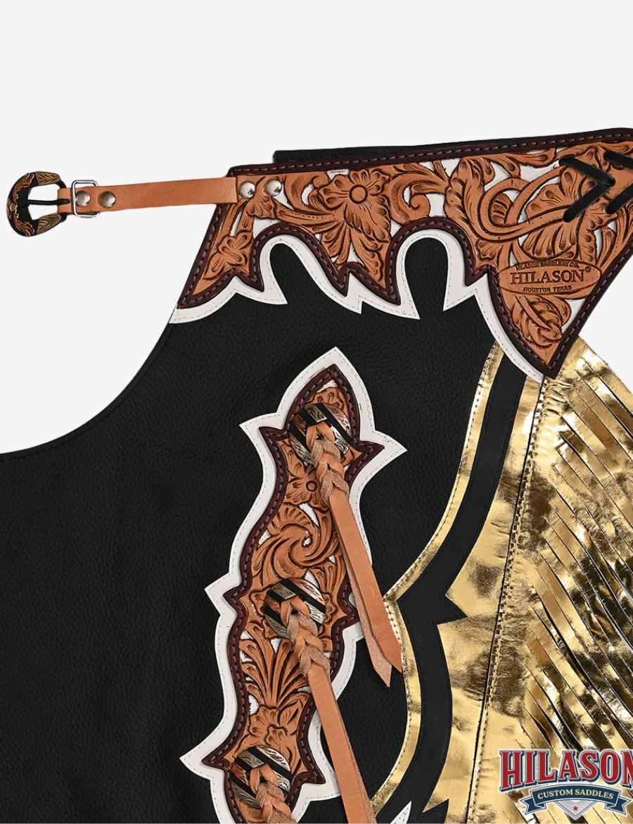 Close-up tooled yoke detail on black rodeo chaps with floral carving and metallic gold fringe accents.