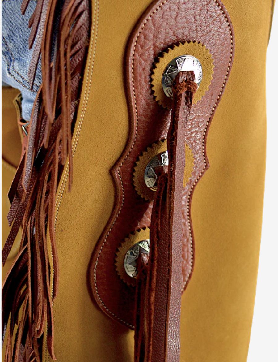 Close tooling detail of golden suede concho chinks chaps with stitched leather accents, conchos, and suede texture.