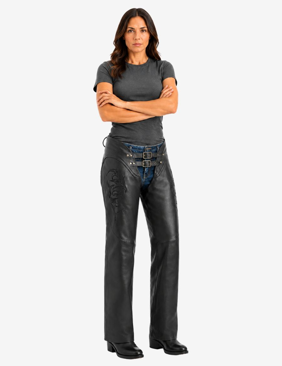 Female model wearing floral embroidered black biker chaps over denim jeans with double buckle belt, styled in a confident motorcycle-ready stance.